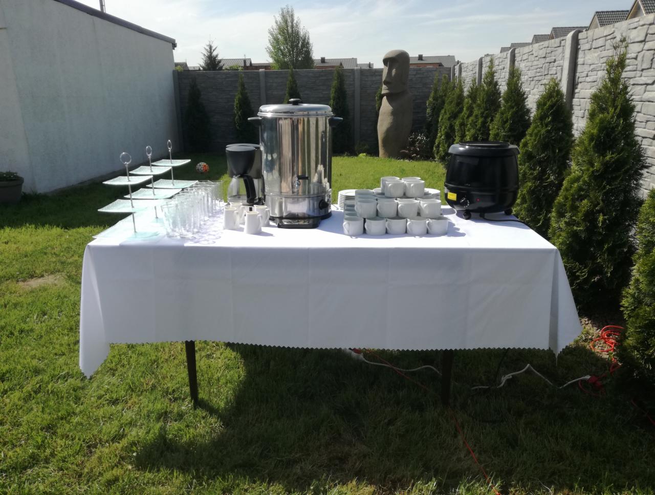 Stół z białym obrusem na trawie, zastawiony naczyniami, szklankami, dyspenserem do kawy i podgrzewaczem do zupy. W tle rzeźba głowy z Wyspy Wielkanocnej i tuje.