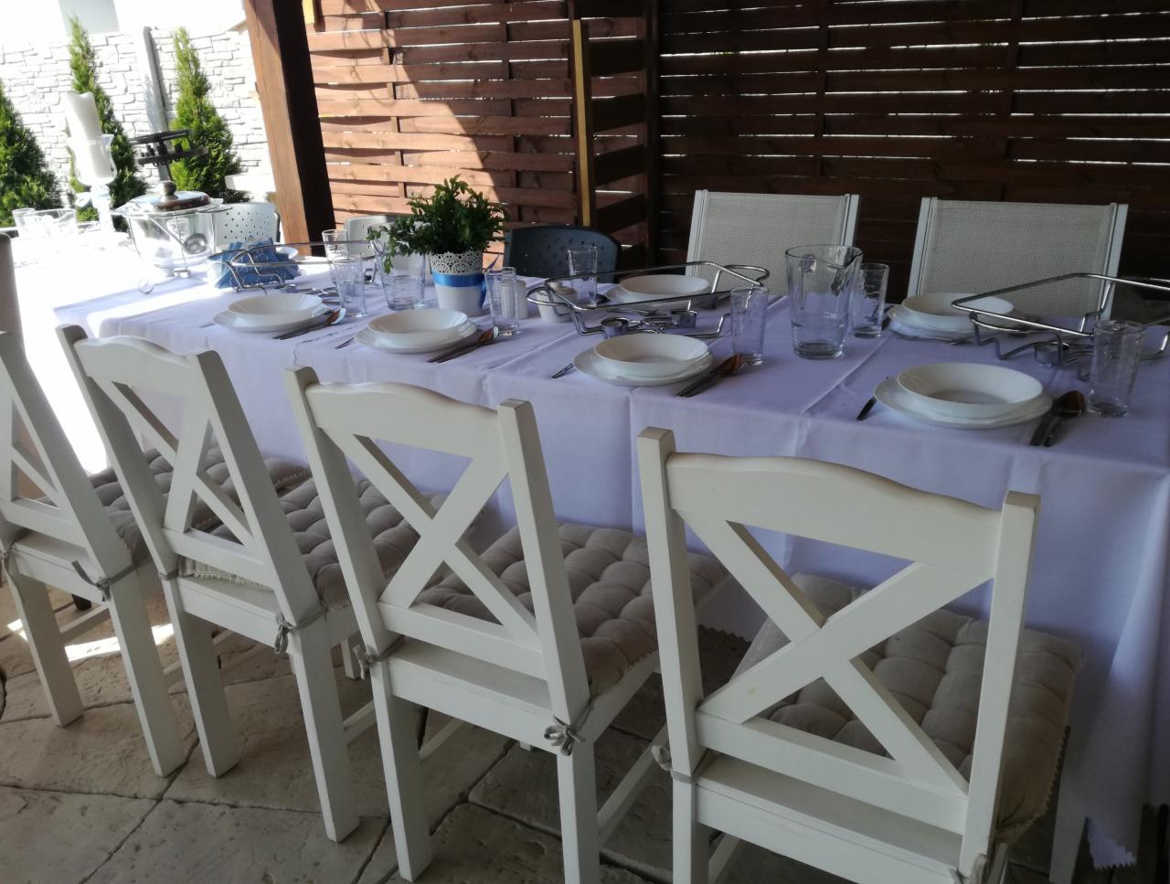 Uroczysty stół nakryty białym obrusem z zastawą dla gości, ustawiony na zewnątrz pod drewnianą pergolą, z białymi krzesłami z poduszkami.