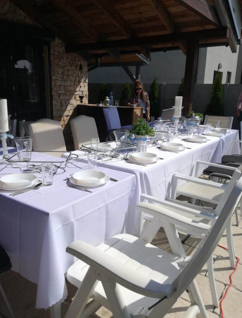 Uroczysty stół z białym obrusem, nakryty zastawą, przygotowany pod zadaszeniem na zewnątrz, z widoczną obsługą w tle.