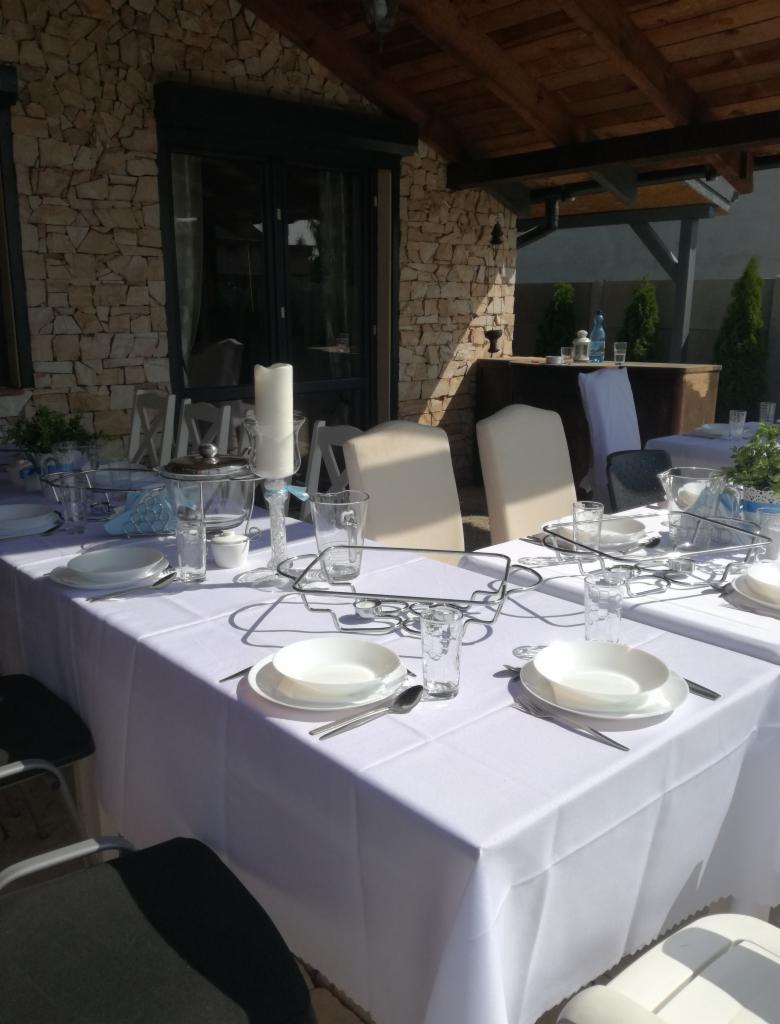 Uroczysty stół z białym obrusem, zastawą i dekoracjami pod zadaszeniem z kamienną ścianą w tle.