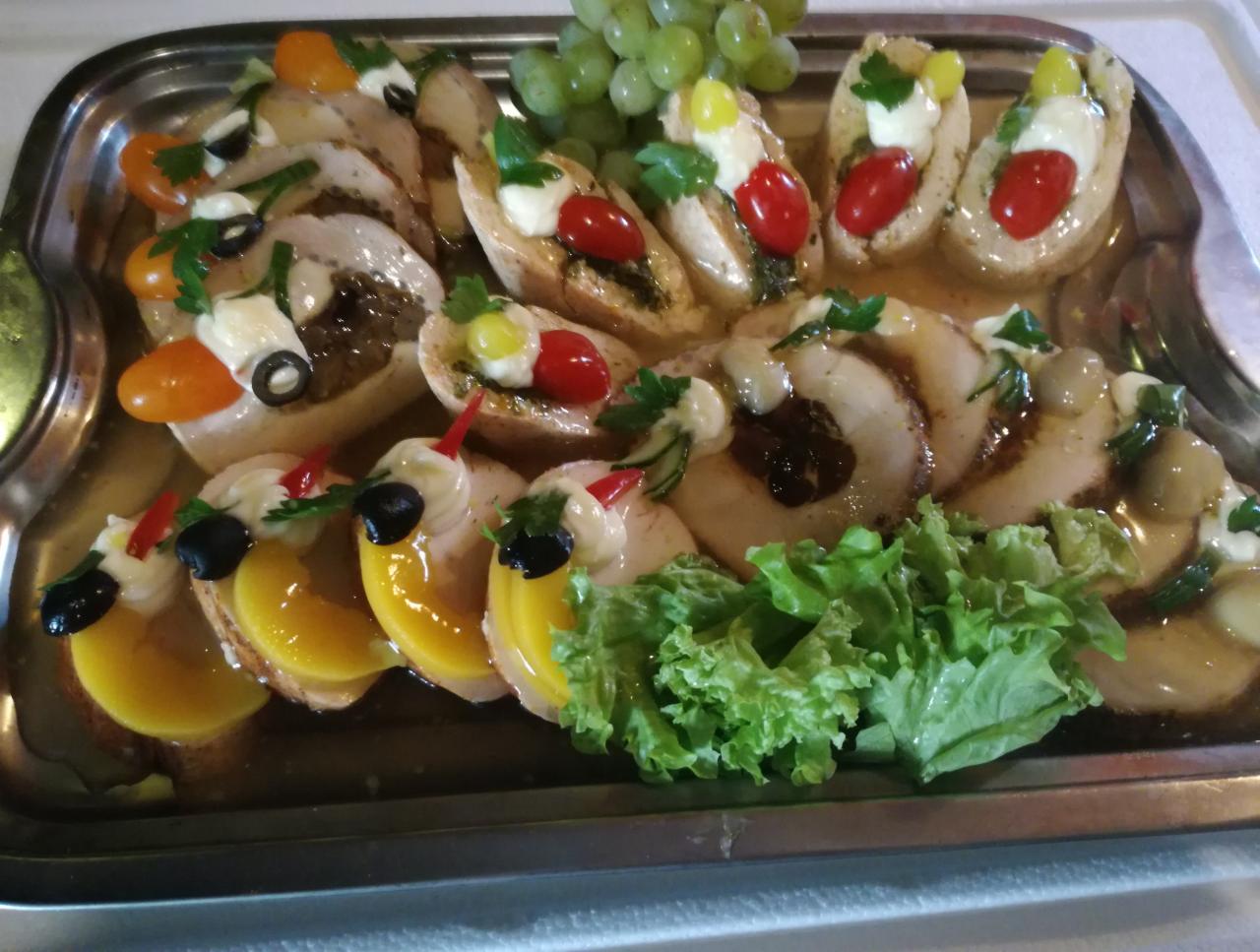 Dekoracyjna taca z przekąskami: roladki mięsne z sosem, grzanki z pomidorkami cherry i pesto, grzanki z brzoskwiniami i oliwkami, winogrona, sałata, całość na srebrnej tacy.