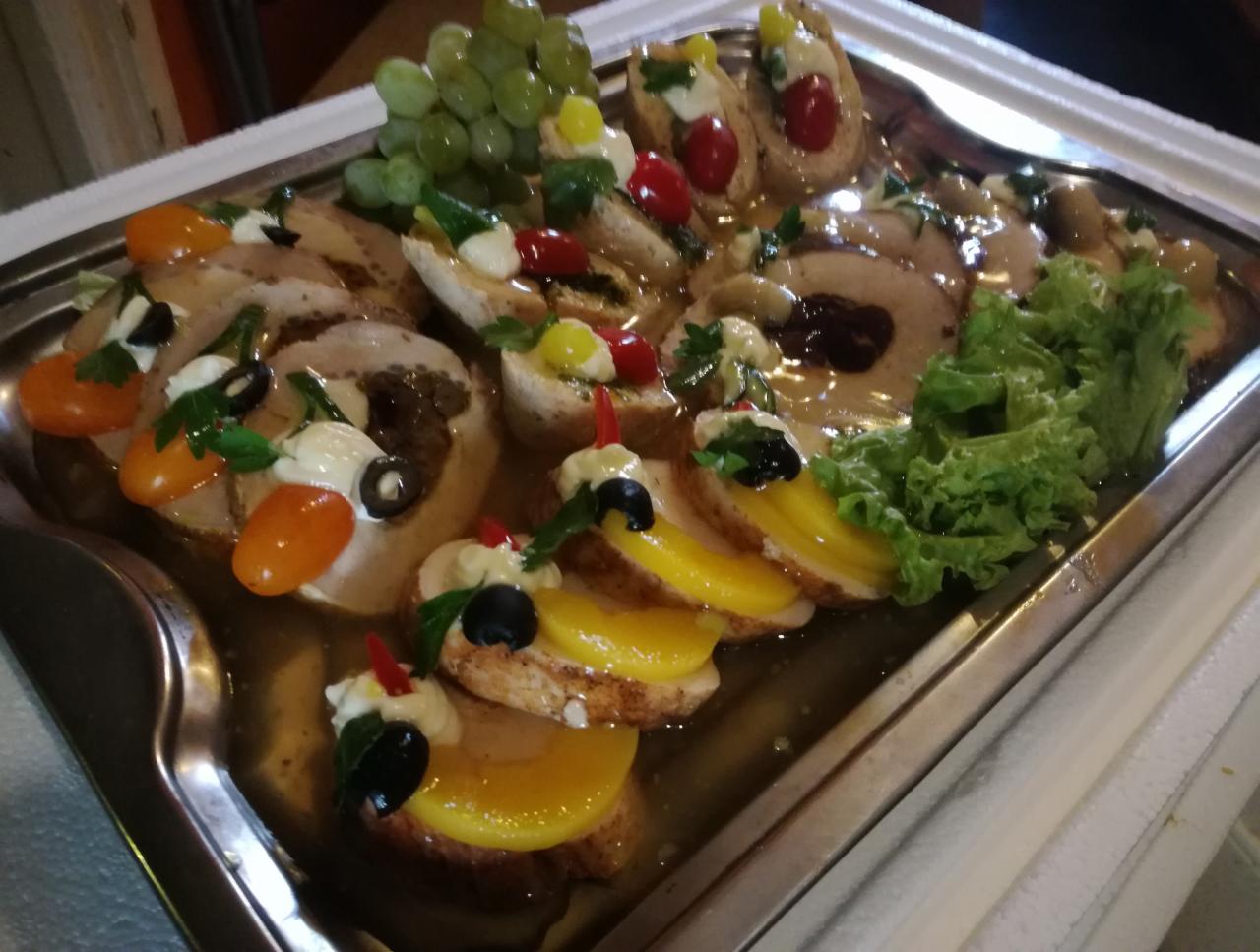 Srebrna taca z roladkami mięsnymi w sosie, udekorowanymi owocami, warzywami i ziołami, tworzącymi apetyczną kompozycję na imprezę.