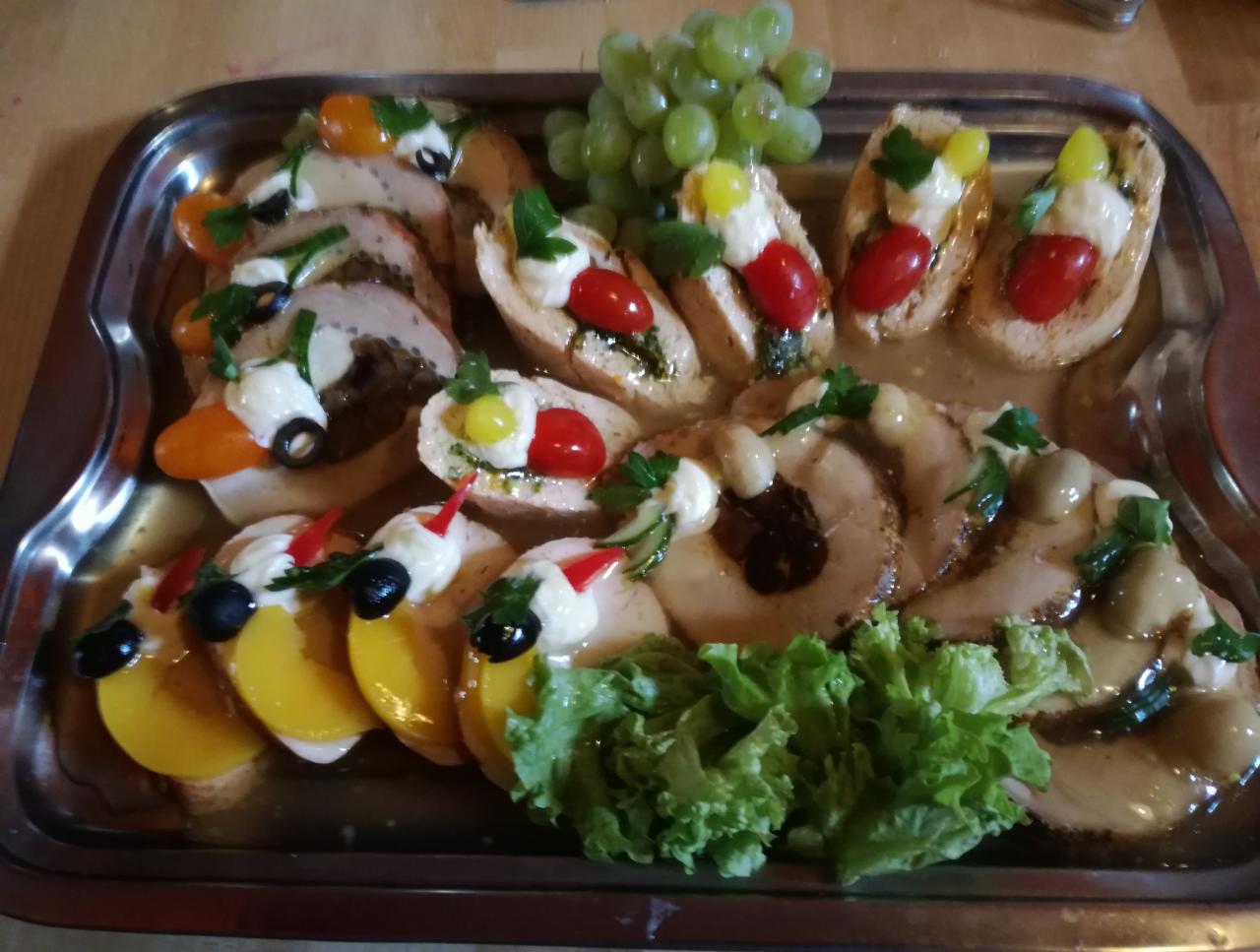 Dekoracyjna taca z różnorodnymi przekąskami: roladki mięsne z sosem, bruschetty z pomidorami i ziołami, plastry mięsa z brzoskwiniami i oliwkami, całość udekorowana świeżymi winogronami i natką...