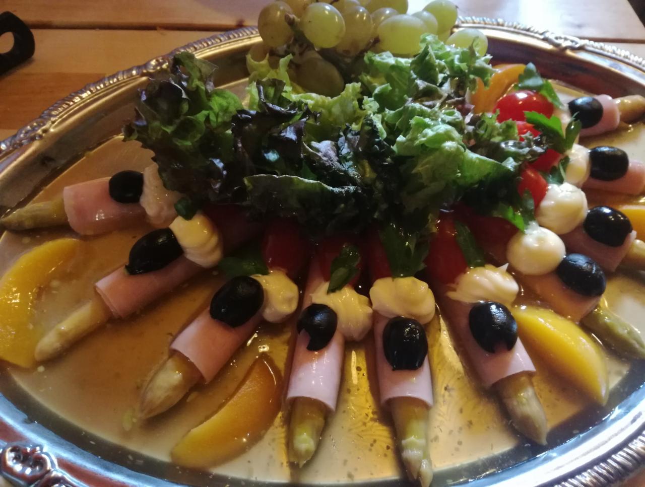 Elegancko podane szparagi owinięte szynką z dodatkiem oliwek, pomidorów, sałaty i winogron na srebrnej tacy, polane sosem.