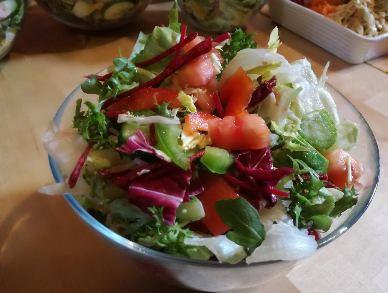 Szklana misa wypełniona kolorową sałatką warzywną z pomidorami, ogórkami, różnymi rodzajami sałat i czerwoną kapustą, widoczna z bliska na drewnianym stole.