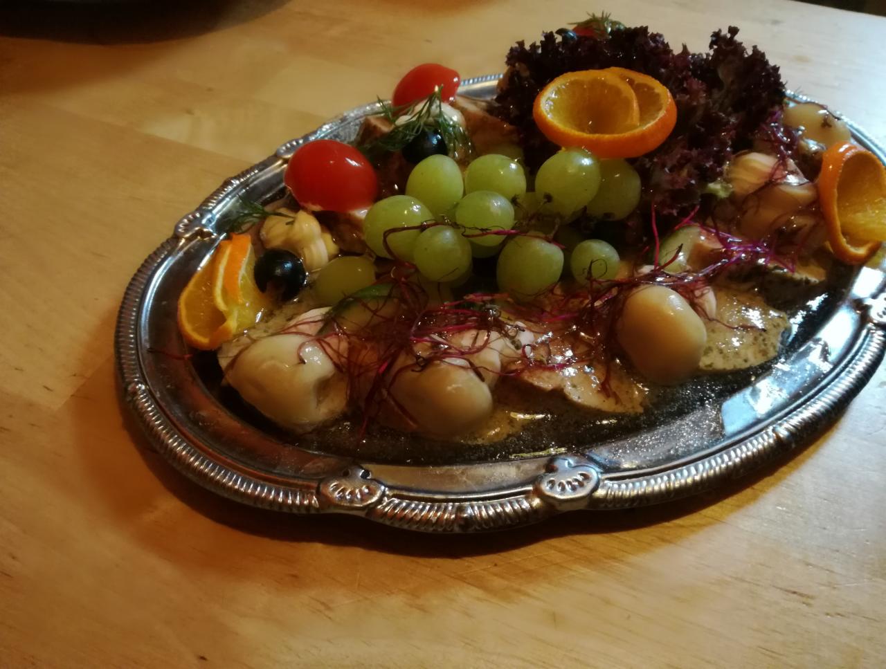 Dekoracyjna srebrna patera z rybą w galarecie, winogronami, pomidorkami koktajlowymi, oliwkami, sałatą i pomarańczą na drewnianym stole.