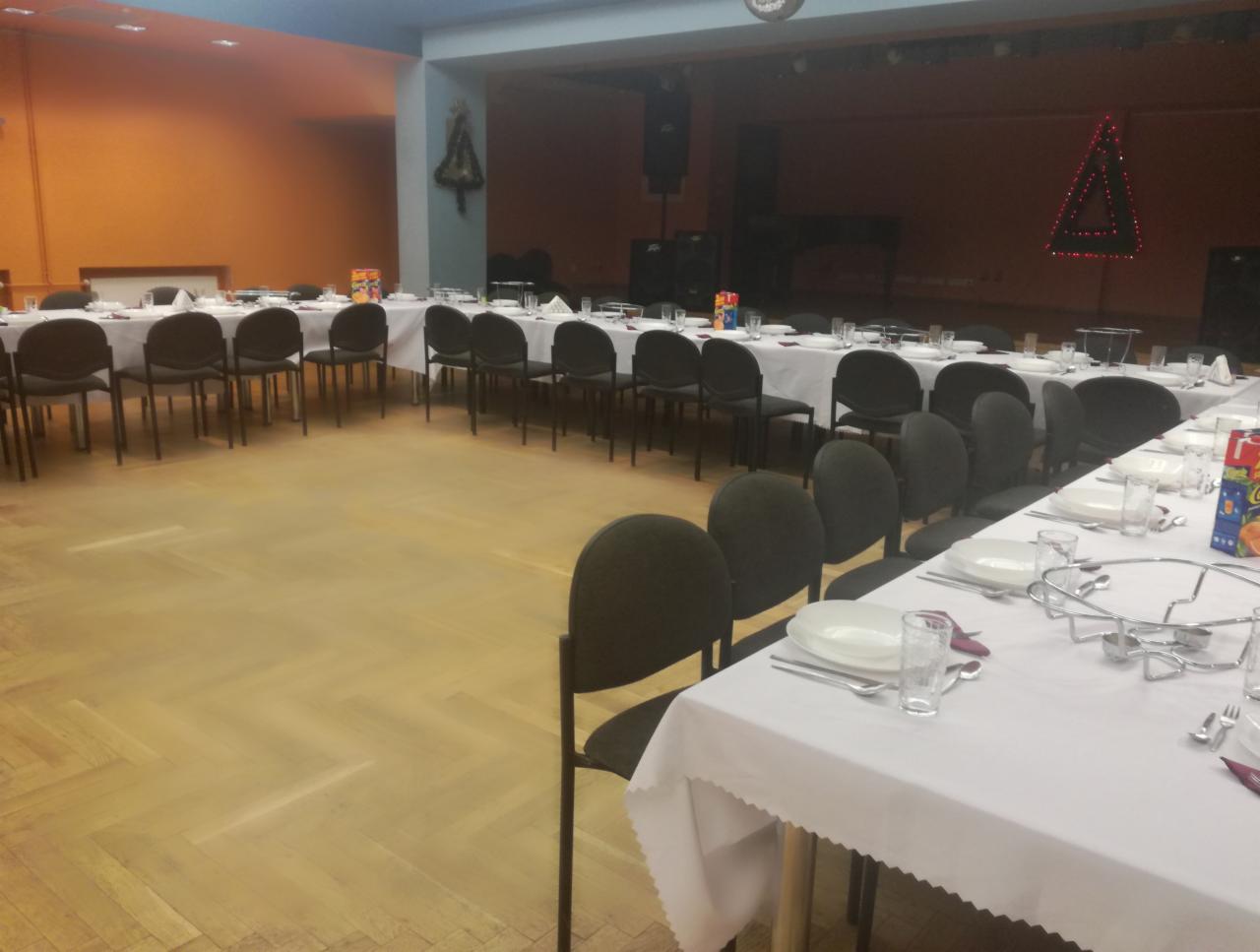 Sala bankietowa z długimi stołami nakrytymi białymi obrusami, zastawionymi talerzami, sztućcami i szklankami, przygotowana na uroczystość. Dekoracje świąteczne w tle, w tym choinka z lampek.