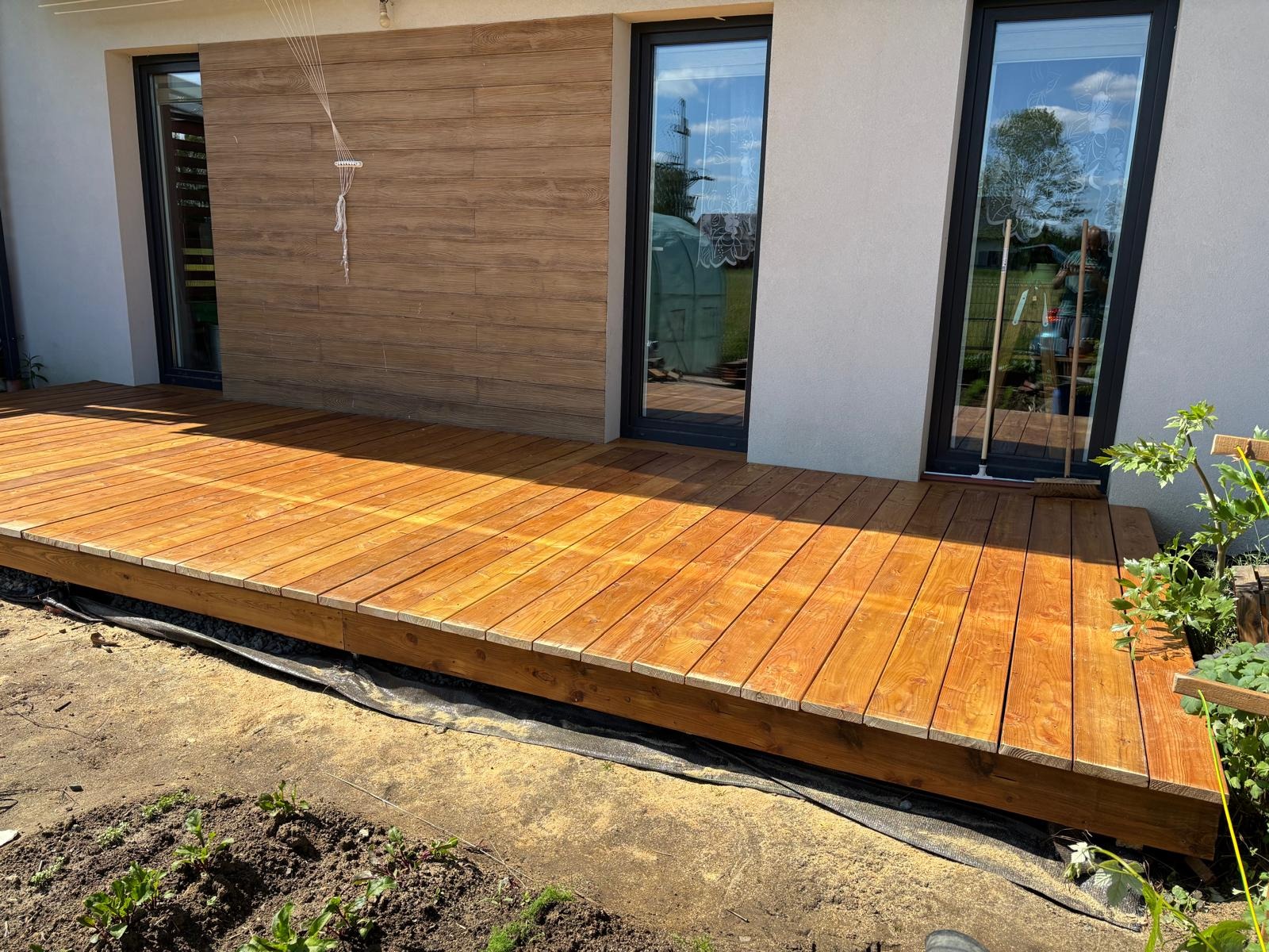 Nowoczesny taras z drewna o ciepłym odcieniu, przylegający do budynku z dużymi oknami. Widoczne deski tarasowe i fragment ogrodu z młodymi roślinami.