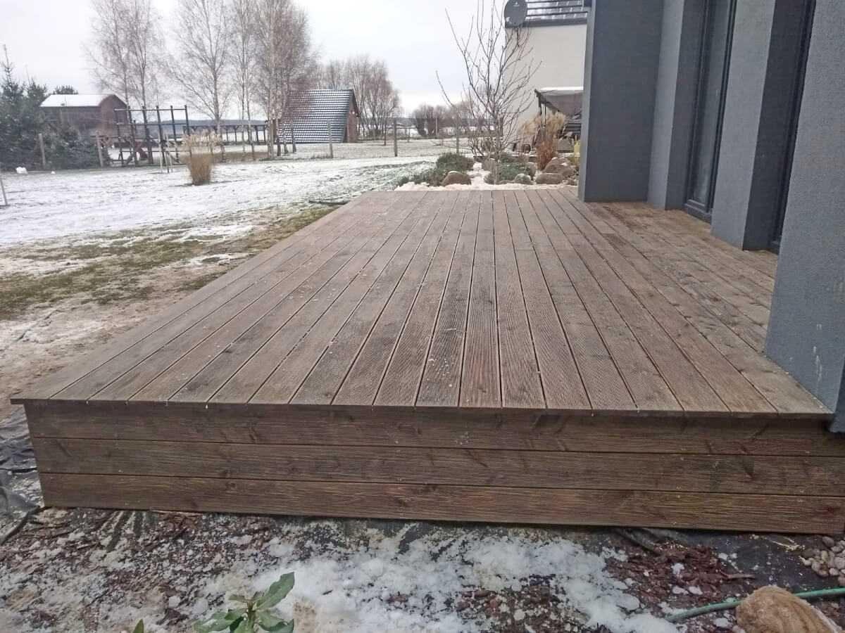 Nowoczesny taras drewniany z desek w odcieniach brązu, przylegający do szarego budynku. W tle zimowy krajobraz z drzewami i domami.