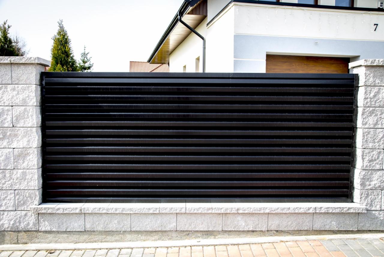 Ciemny, poziomy płot lamelowy z kamiennymi słupkami na tle domu z numerem 7.