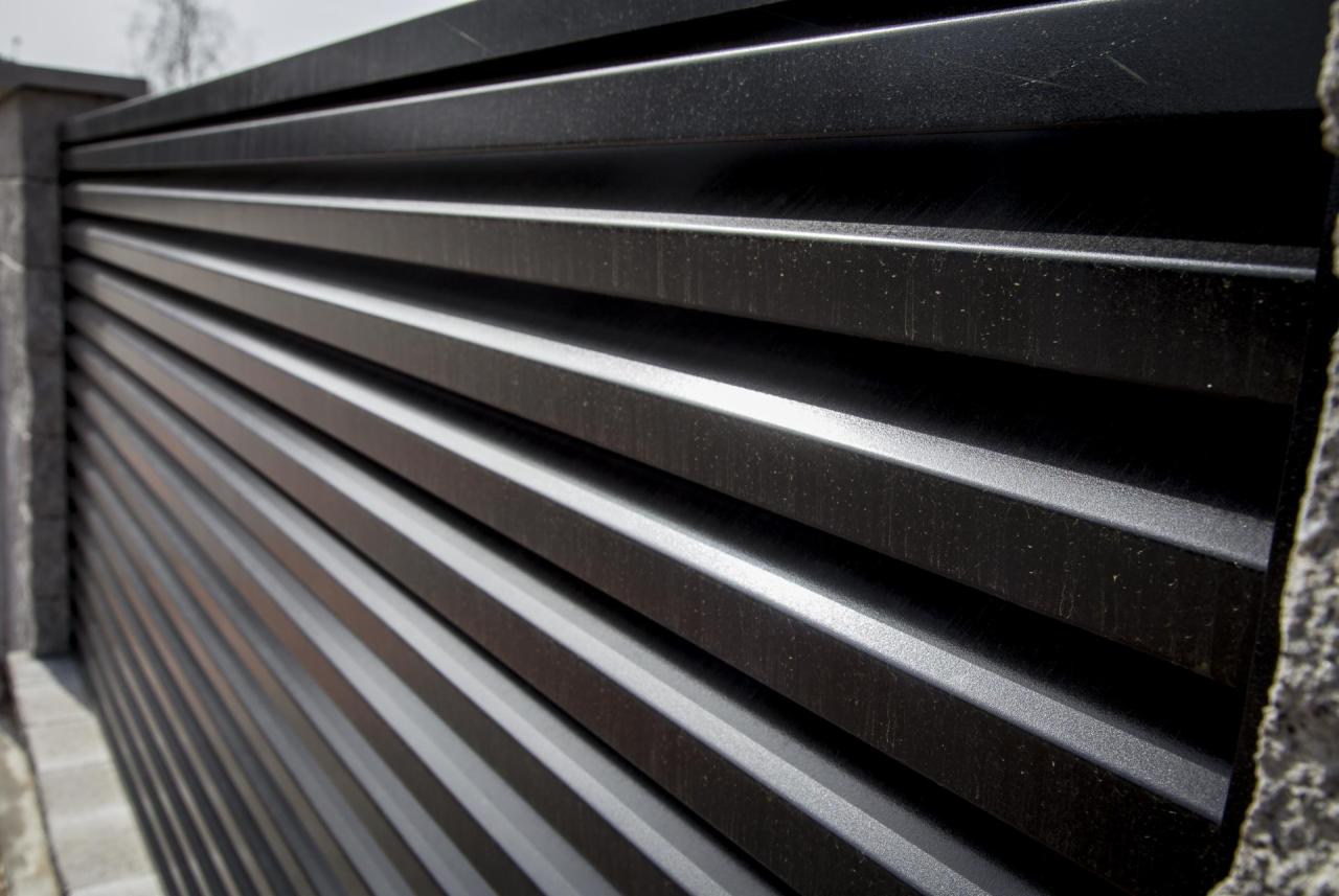 Fragment czarnego, poziomego ogrodzenia z metalowych profili, widok z boku, z betonowym słupkiem po lewej i prawej stronie.