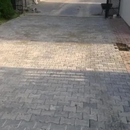 Częściowo ułożona szara kostka brukowa typu 'I' na posesji, widoczna ubijarka wibracyjna i przygotowany fragment pod ułożenie kolejnej partii kostki.