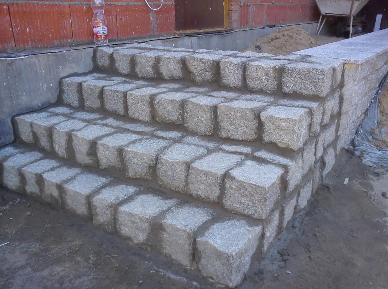 Granitowe schody wejściowe do budynku w trakcie budowy, z widoczną butelką wody na szczycie i taczką w tle.