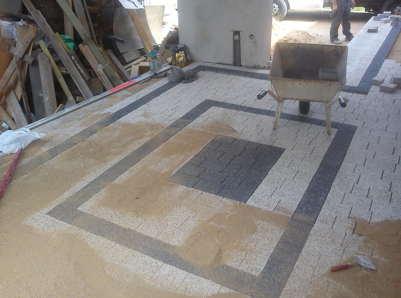 Fragment układanej nawierzchni z kostki brukowej w geometryczny wzór z piaskiem, widoczna taczka, narzędzia i materiały budowlane w tle.