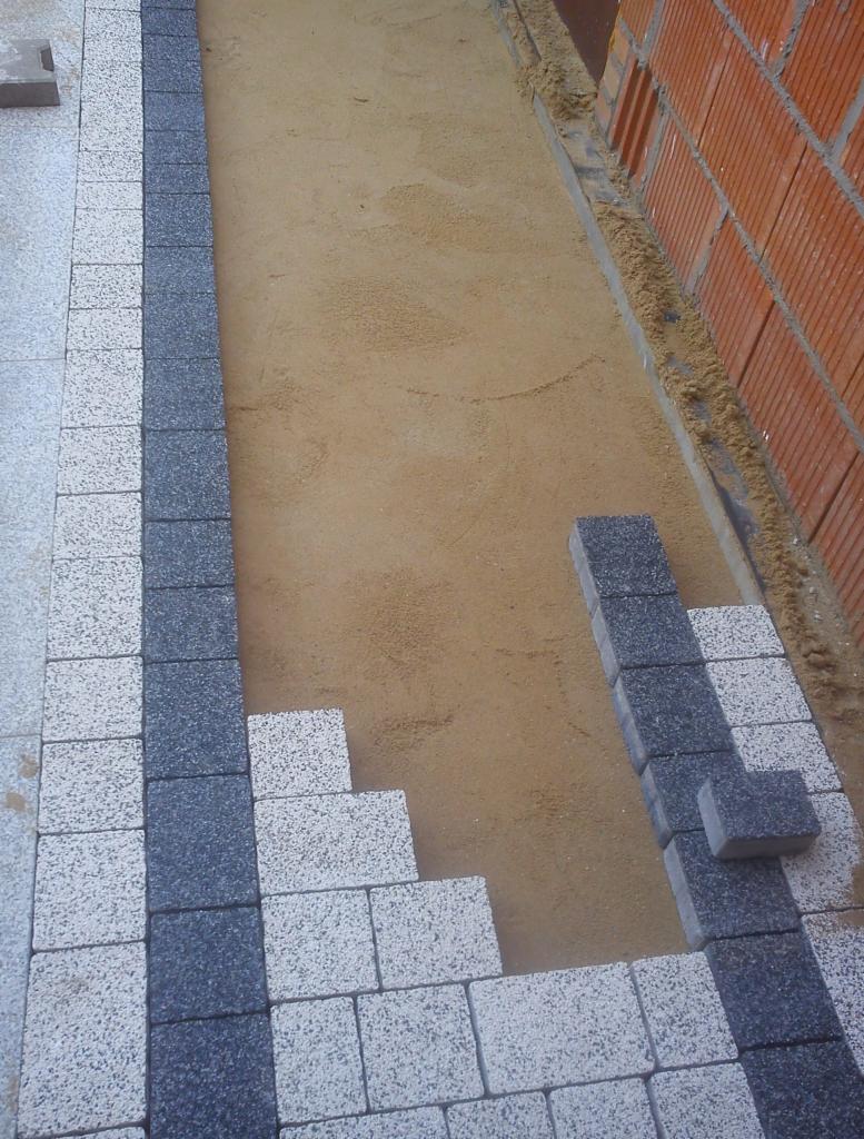 Fragment układanej nawierzchni z kostki brukowej w kolorach szarości, widoczny piasek podkładowy i fragment ściany budynku