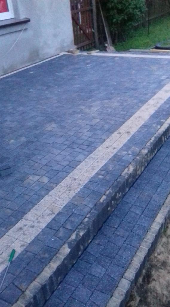 Ułożona, grafitowa kostka brukowa z jasnym pasem wzdłuż krawędzi, prowadząca do drzwi wejściowych, widoczne ślady po pracach brukarskich.