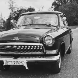 Czarno-białe zdjęcie klasycznego samochodu z tabliczką 'Nowożeńcy' na przednim zderzaku, w tle para młoda idąca drogą.