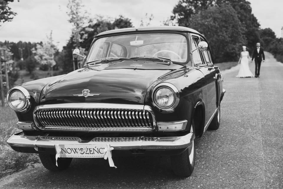 Czarno-białe zdjęcie klasycznego samochodu z tabliczką 'Nowożeńcy' na przednim zderzaku, w tle para młoda idąca drogą.
