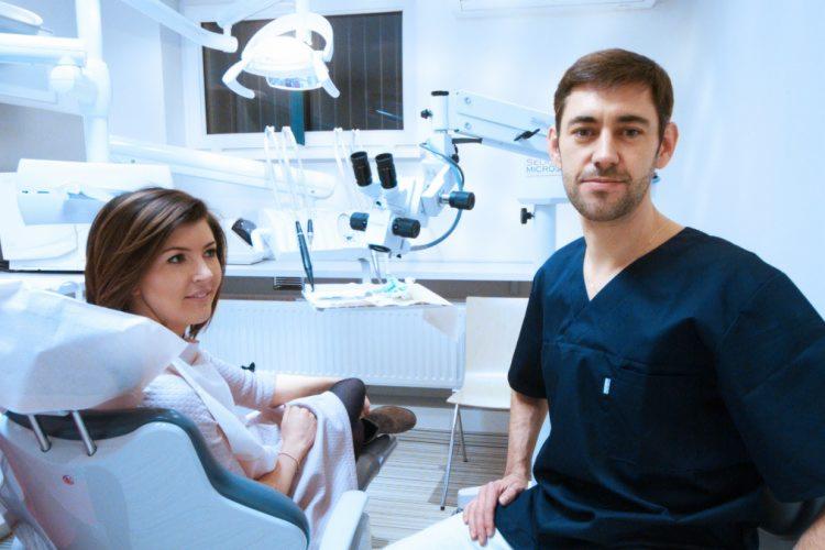 Pacjentka siedzi na fotelu dentystycznym, obok stoi lekarz w granatowym uniformie, w tle widoczny mikroskop zabiegowy i lampa oświetleniowa.