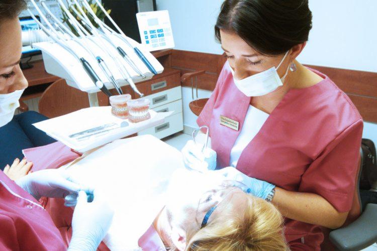 Dentystka w różowym uniformie i masce wykonuje zabieg na pacjencie leżącym na fotelu dentystycznym, widoczne narzędzia, model szczęki i aparat sterujący