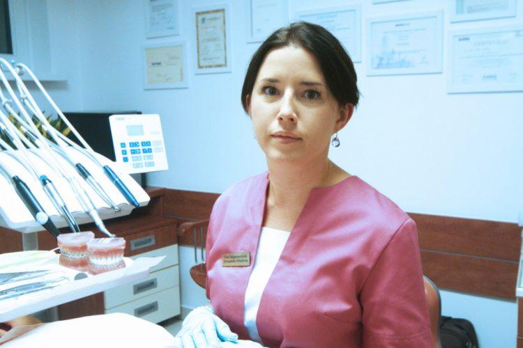 Portret kobiety w różowym uniformie stomatologicznym na tle gabinetu z modelami szczęk i narzędziami dentystycznymi.