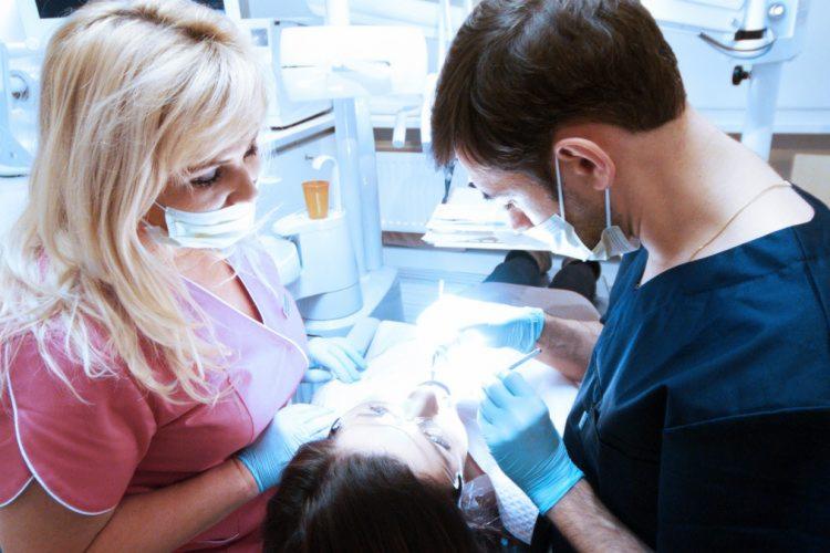 Pacjentka podczas zabiegu w gabinecie, dwóch lekarzy w maskach i rękawiczkach pochyla się nad nią, używając narzędzi dentystycznych.