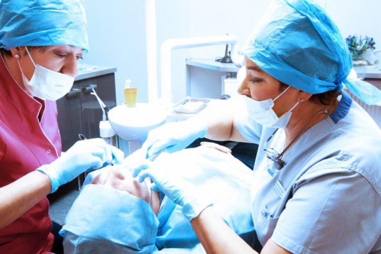 Dwaj lekarze w strojach medycznych pochyleni nad pacjentem podczas zabiegu stomatologicznym w jasnym gabinecie.