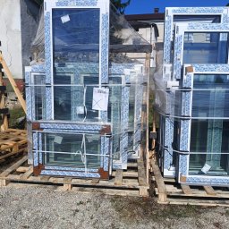 P.P.H.U Lukas Okna - Stos okien PCV na paletach, zabezpieczonych folią, z widocznym profilem w kolorze białym i brązowym, na zewnątrz budynku w słoneczny dzień.