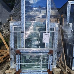 P.P.H.U Lukas Okna - Paleta z zapakowanymi oknami o białych i brązowych ramach, zabezpieczonymi folią stretch, na zewnątrz budynku w słoneczny dzień. Widoczny budynek w tle.