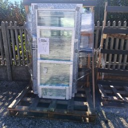 P.P.H.U Lukas Okna - Nowe, białe okna PCV, zabezpieczone folią, ułożone na drewnianej palecie na zewnątrz, gotowe do montażu. Widoczne etykiety producenta.