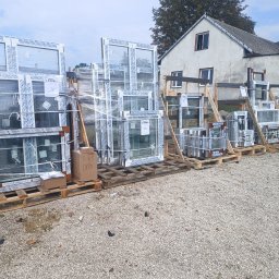 P.P.H.U Lukas Okna - Okna PCV Salamander, przygotowane do montażu w Pińczowie, zabezpieczone folią na paletach. Widoczne kartony z oznaczeniami i drewniane podpory.