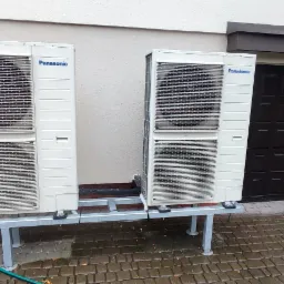 Dwie jednostki zewnętrzne klimatyzacji Panasonic zamontowane na metalowej konstrukcji przed budynkiem z brązowymi drzwiami garażowymi i oknem, widoczny fragment zielonego węża.