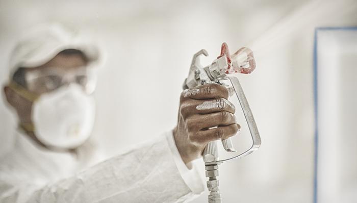 Pracownik w kombinezonie ochronnym i masce maluje ścianę natryskowo, trzymając w dłoni pistolet do malowania; widoczne ślady farby na dłoni.