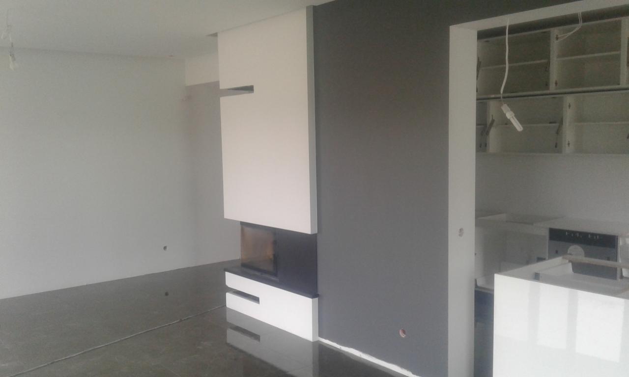 Wnętrze w trakcie wykańczania: biała zabudowa kominka na tle szarej ściany, widoczny fragment kuchni z niedokończonymi szafkami.