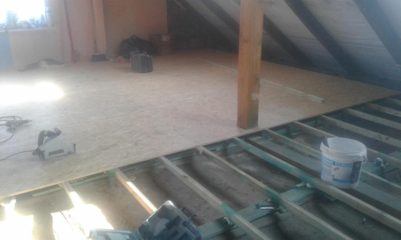 Montaż podłogi z płyt OSB na drewnianej konstrukcji szkieletowej w trakcie prac, widoczna piła elektryczna i wiadro z klejem.