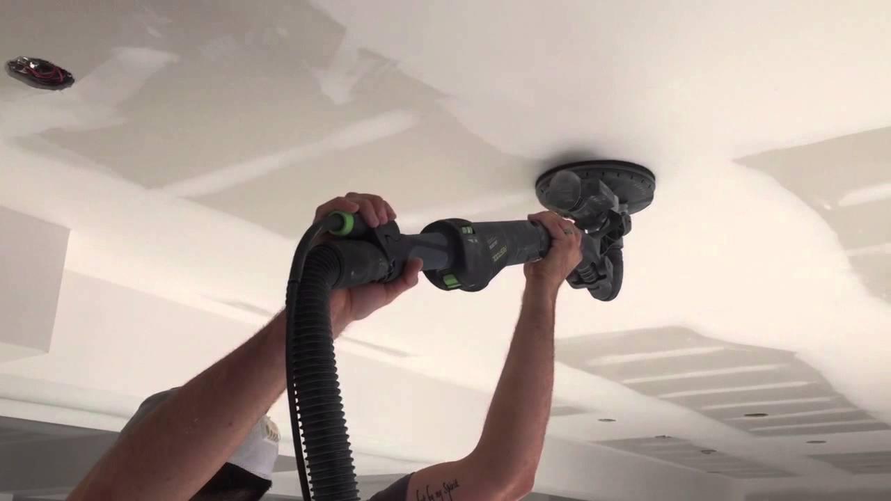 Szlifierka do gładzi gipsowych w trakcie pracy na suficie podwieszanym z płyt kartonowo-gipsowych, widoczne łączenia płyt. Operator w czapce z daszkiem.