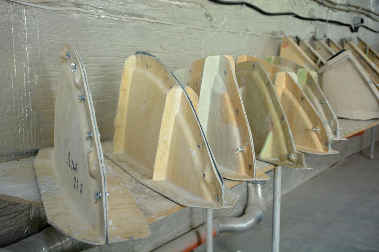 Szereg beżowych, trójkątnych form kompozytowych z przykręconymi śrubami, ustawionych na metalowym regale w warsztacie.