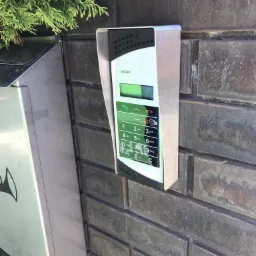 Zewnętrzny panel domofonowy z klawiaturą numeryczną i czytnikiem kart, zamontowany na ceglanym murze obok skrzynki na listy, z zielonymi roślinami w tle.