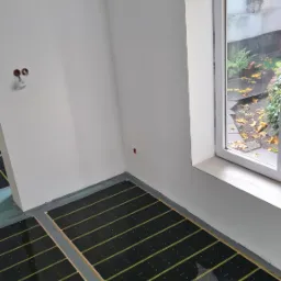 Montaż folii grzewczej na podłodze w jasnym pomieszczeniu z oknem. Widoczne kable elektryczne wystające ze ściany. Układ folii r&oacute;wnoległy do okna.