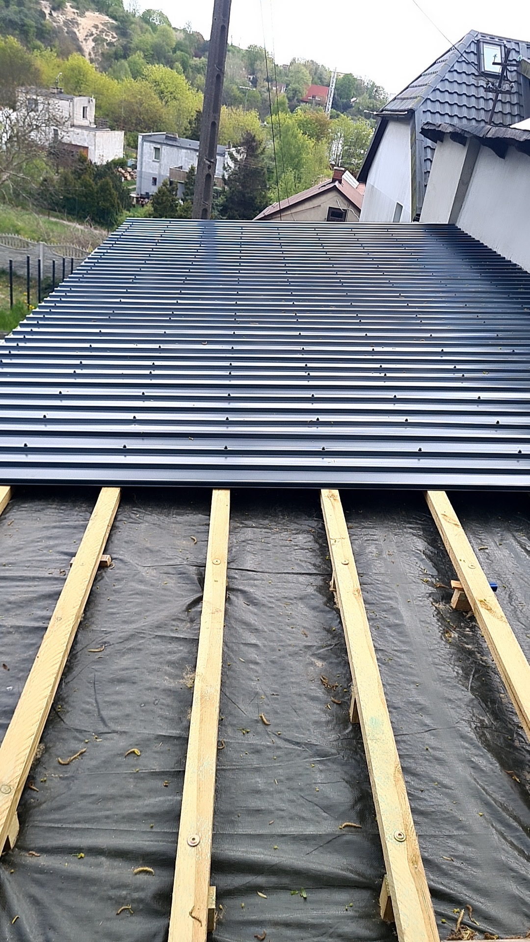 Montaż blachy trapezowej na dachu z widoczną konstrukcją drewnianą i membraną dachową. Widok z góry na budynek w tle.