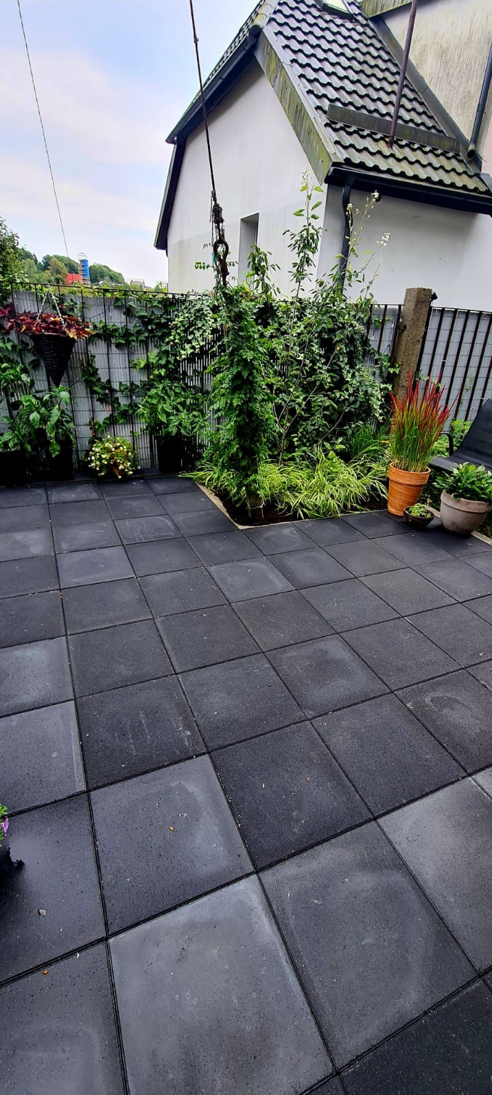 Ułożona, czarna, gumowa kostka na tarasie. W tle zielona ściana z roślin pnących, donice i część budynku z szarą dachówką. Widok na zadbany ogród.
