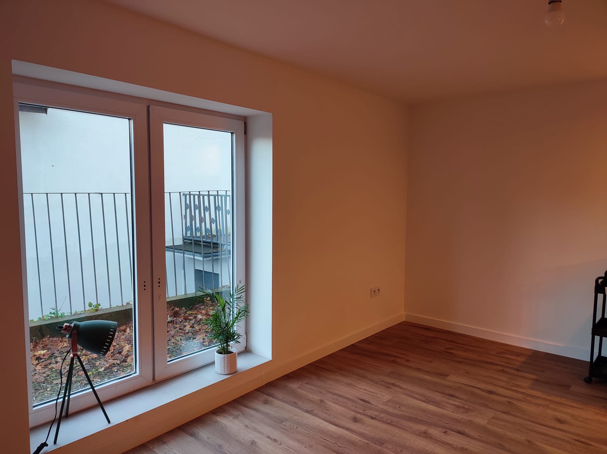 Wnętrze odnowionego pokoju z drewnianą podłogą i świeżo pomalowanymi ścianami w odcieniach bieli. Na parapecie stoi roślina w ozdobnej doniczce i lampka.