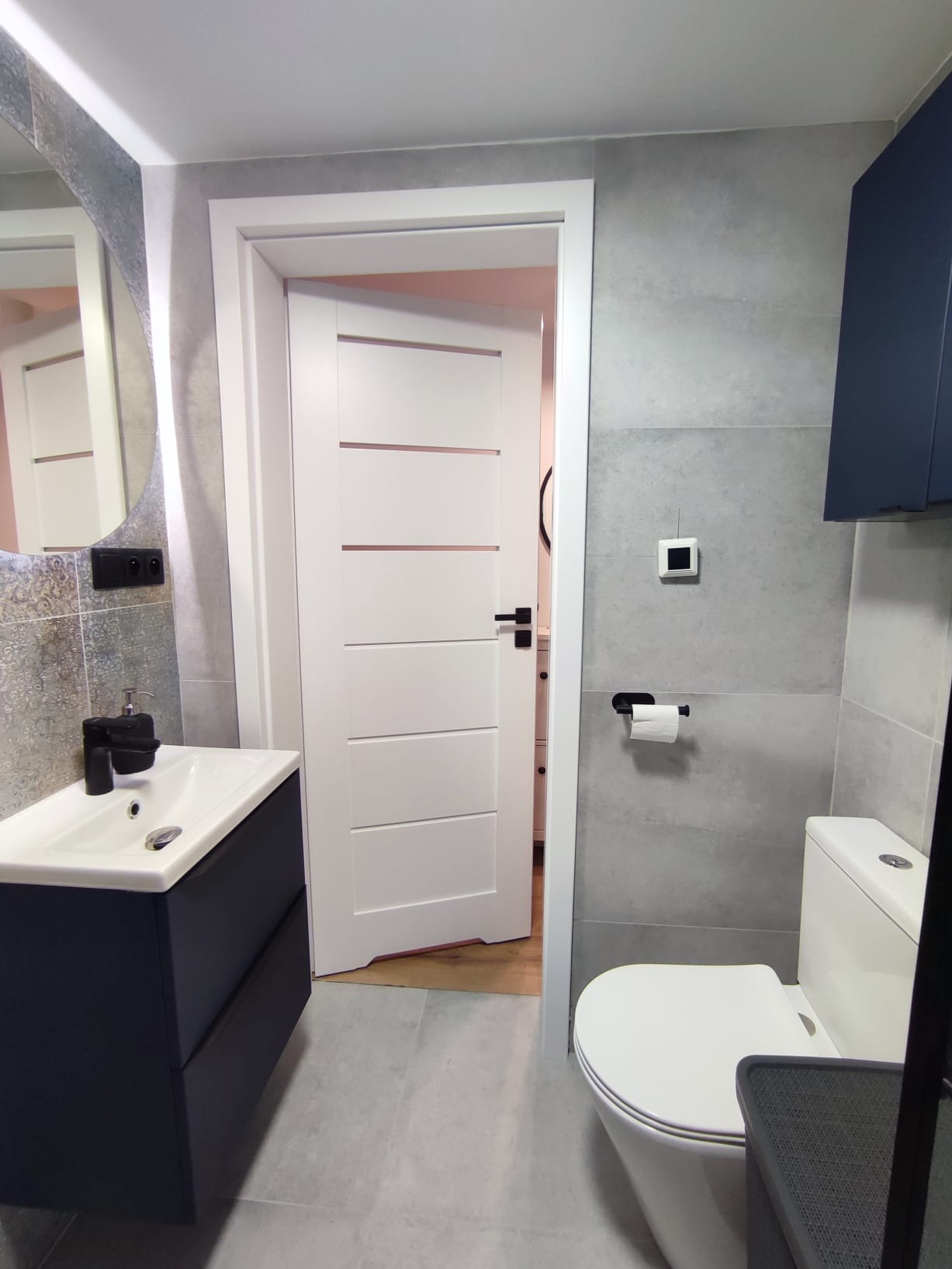 Nowoczesna łazienka z szarymi płytkami, białą armaturą i szafką umywalkową w odcieniach granatu. Widoczna biała, częściowo otwarta, wewnętrzna  stolarka drzwiowa.