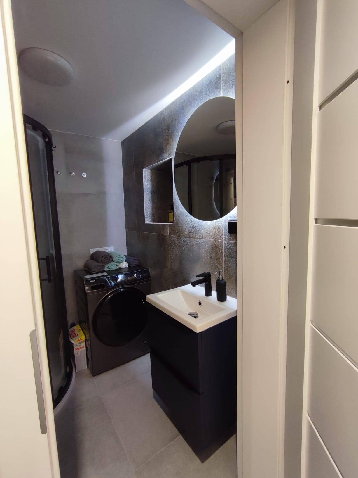 Nowoczesna łazienka z okrągłym lustrem podświetlanym LED, czarną umywalką, kabiną prysznicową i pralką. Szare płytki na ścianach i podłodze.