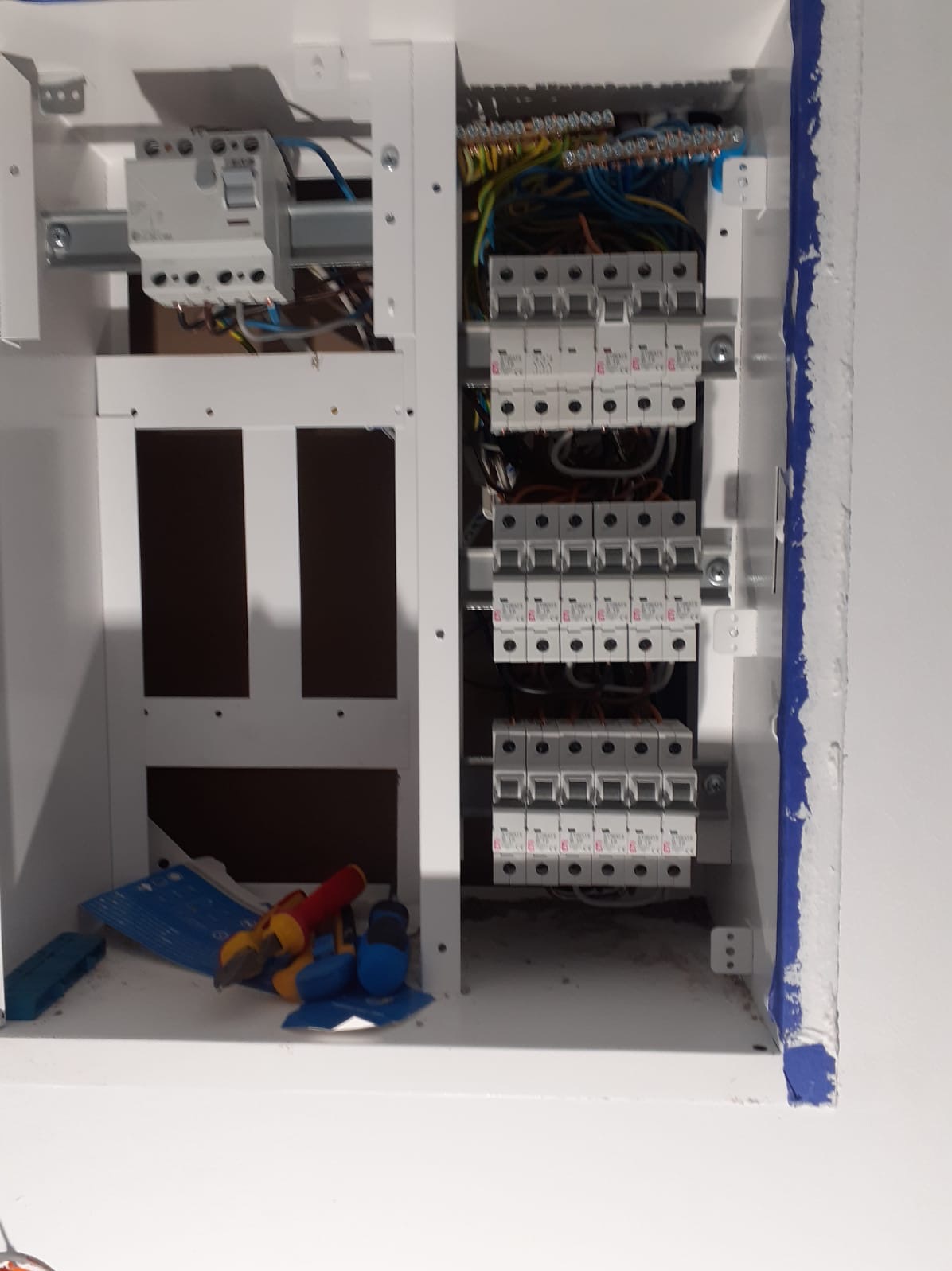 Rozdzielnia elektryczna w białej obudowie z widocznymi bezpiecznikami, przewodami i narzędziami. Świeżo pomalowana ściana z niebieskimi śladami farby.