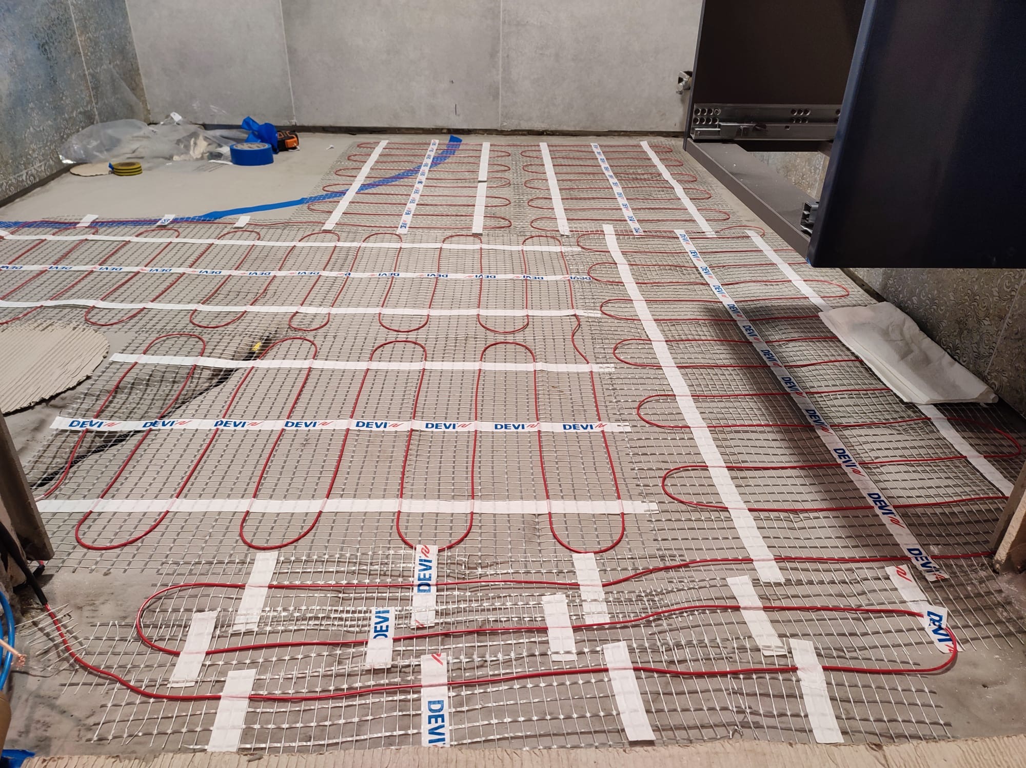 Rozłożona mata grzewcza DEVI z czerwonymi kablami na szarej podłodze w trakcie instalacji ogrzewania podłogowego. Widoczne taśmy mocujące i fragment szafki.