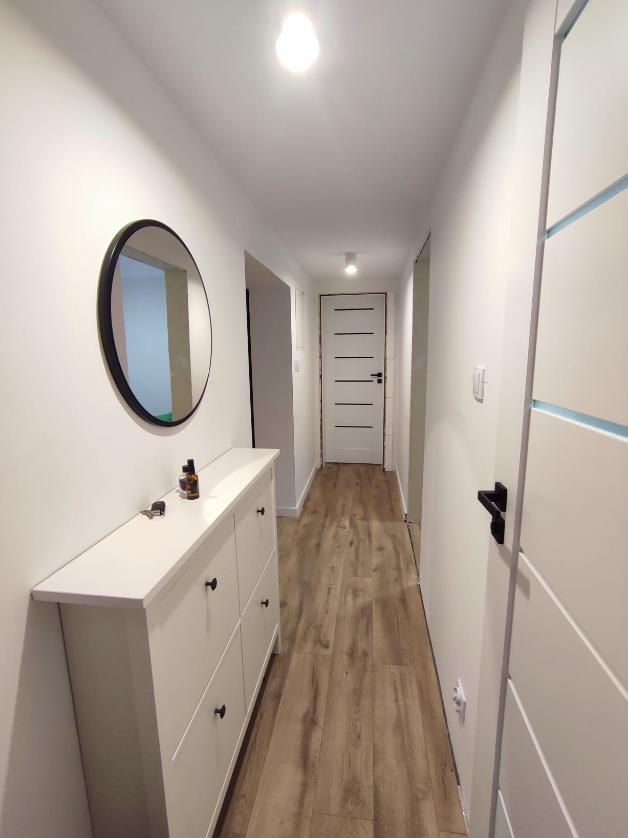 Jasny korytarz po remoncie: białe ściany, drewnopodobna podłoga, biała szafka na buty z okrągłym lustrem. Widoczne drzwi z poziomymi frezami.