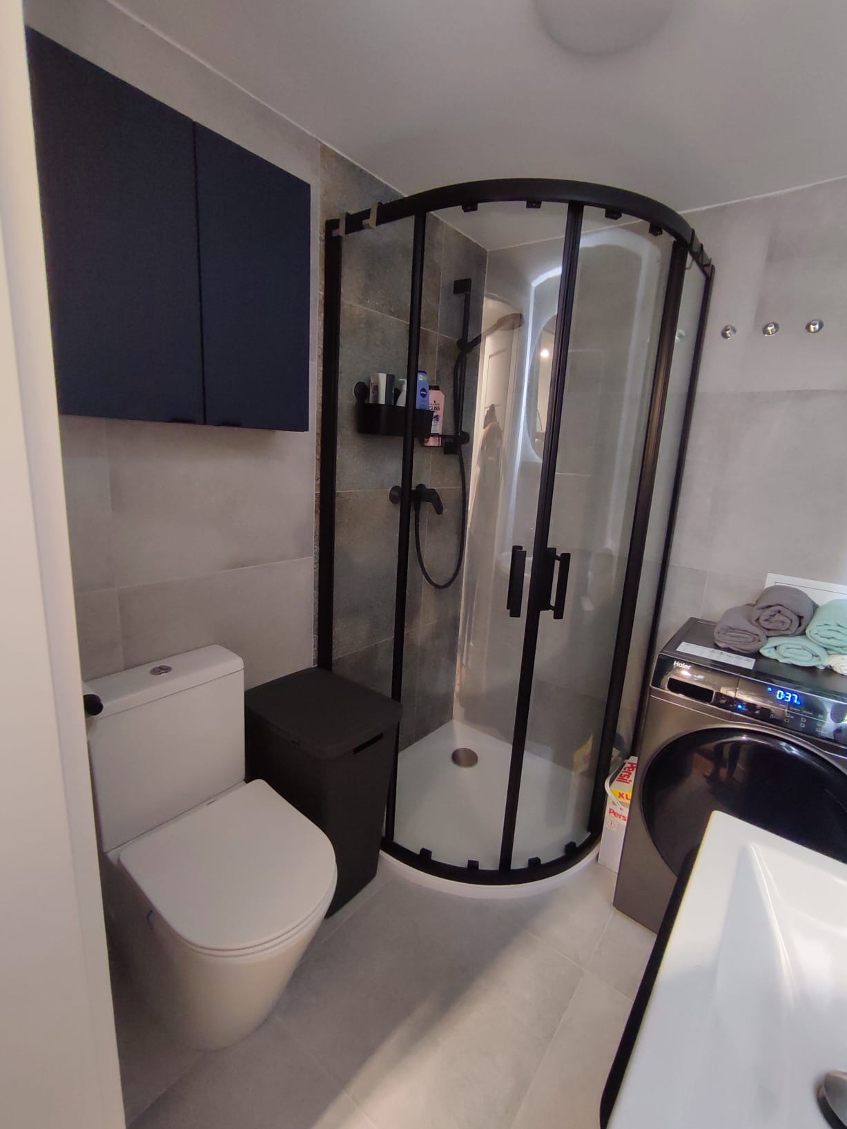 Nowoczesna łazienka z kabiną prysznicową typu walk-in, toaletą, szafką i pralką. Szare płytki, czarne akcenty, minimalistyczny design.