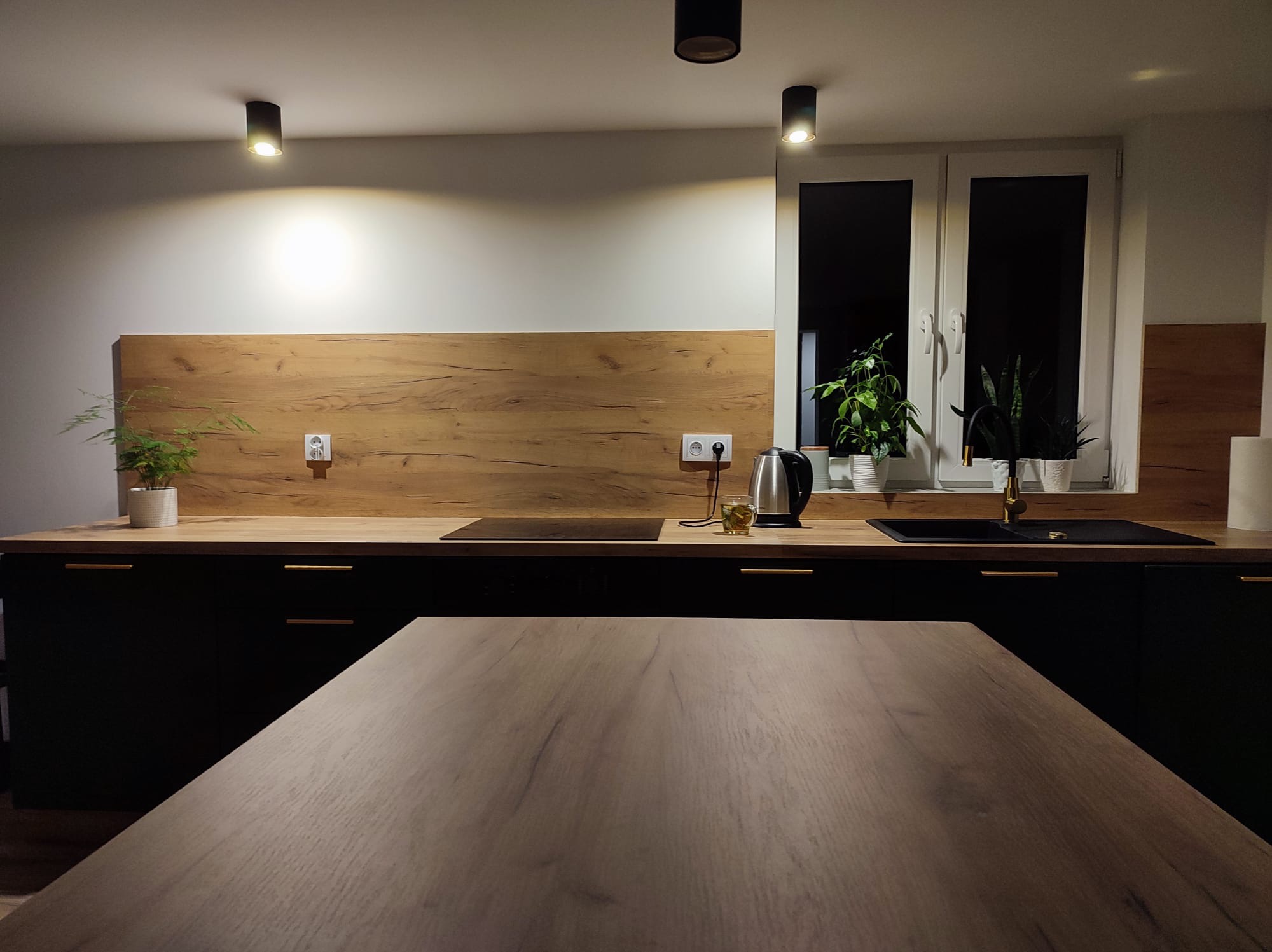 Nowoczesna kuchnia z czarnymi szafkami, drewnianym blatem i panelem, oświetleniem punktowym i roślinami w oknie. Stół na pierwszym planie. Ciepłe światło.