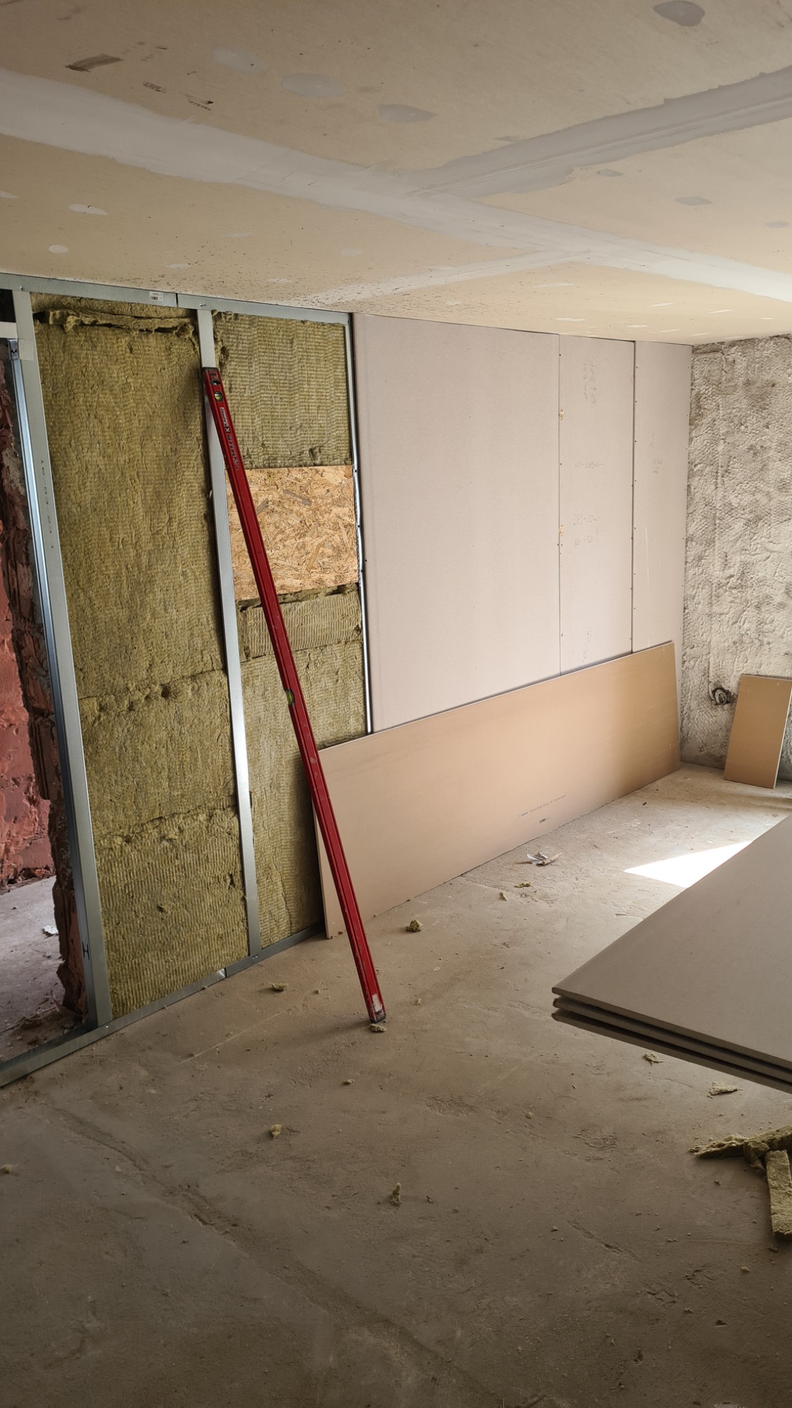 Wnętrze w trakcie remontu: metalowa konstrukcja ściany z izolacją, płyty gipsowo-kartonowe oparte o ścianę, poziomica w pionie, surowy beton na podłodze.